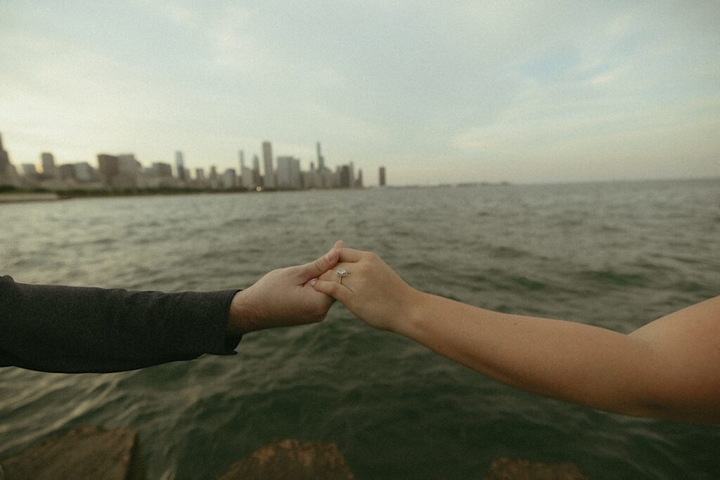 A bride and groom hold hands showcasing the brides princess cut engagement ring overlooking Lake Michigan. 