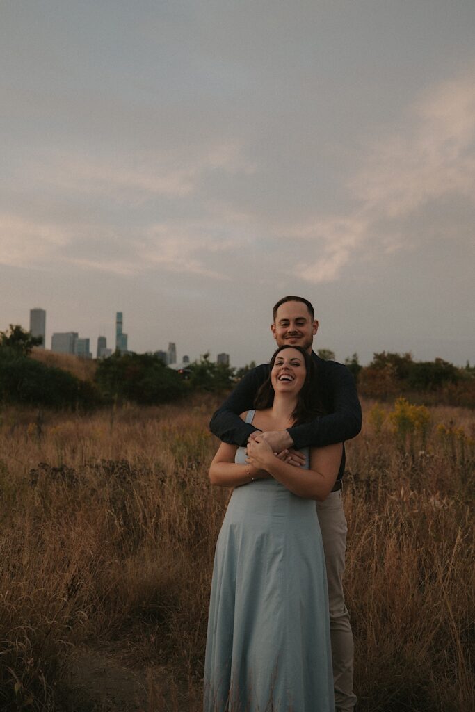 A bride and groom stand holding one another and laughing at the photographer taking their photo during their Chicago engagement session. 