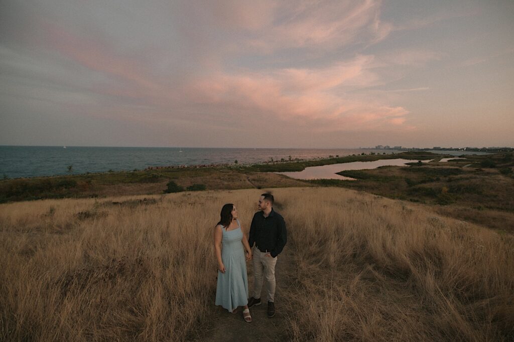 A couple kisses during sunset at an engagement session in Chicago in a field north of the city overlooking lake Michigan and a smaller body of water. 