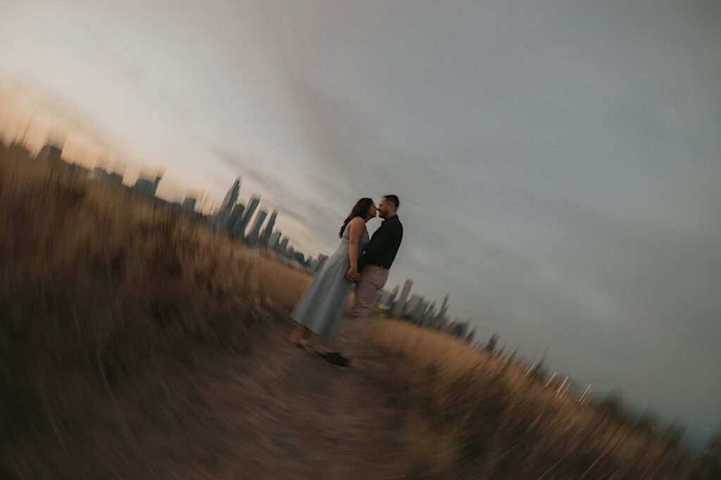 A couple kisses during sunset in a field overlooking the city.  You can see the Chicago skyline behind them and the photographer twisted the photo to make it purposely blurry. 