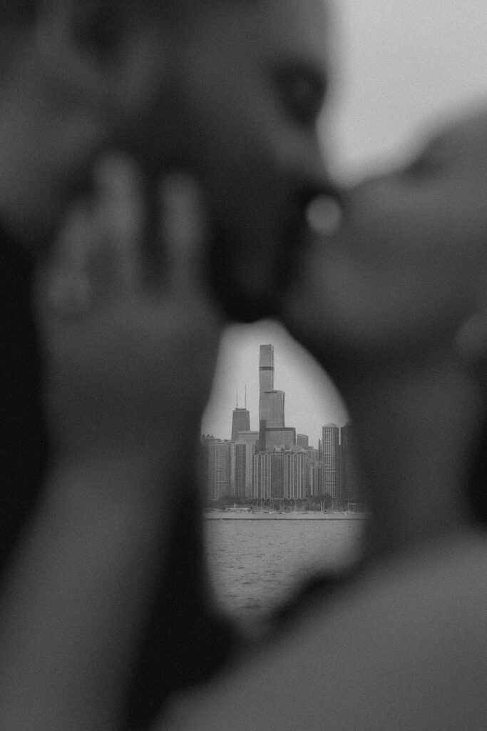 A couple kisses and between their faces you can see the Chicago Skyline in focus behind them.  