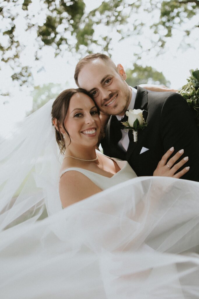 A bride and groom smile for a portrait framed by their veil during their wedding at their Chicago suburbs wedding venue. 