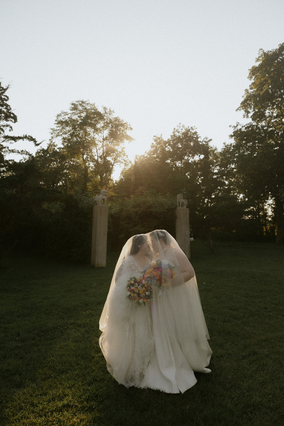 two brides stand under a veil side by side during golden hour at Allerton Park for their elopement